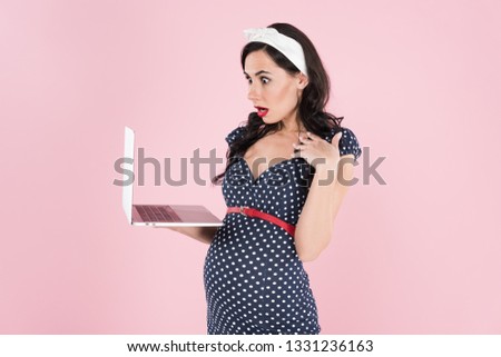 Surprised pregnant woman in dotted dress looking at laptop screen isolated on pink