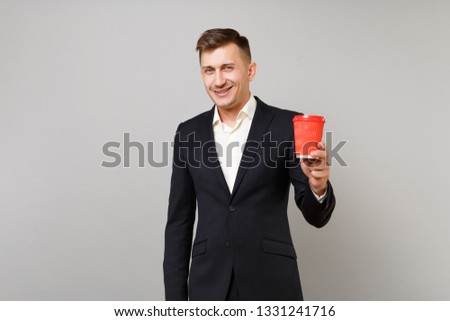 Smiling young business man in classic black suit, shirt holding paper cup with coffee or tea isolated on grey wall background in studio. Achievement career wealth business concept. Mock up copy space