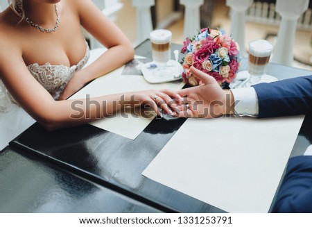 The groom holds the bride's hand in the cafe. Cups with cappuccino and bouquet from red rose.