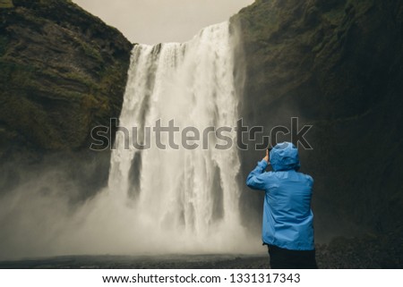 man in blue jacket taking a picture of skogafoss Iceland most famous waterfall on his phone 