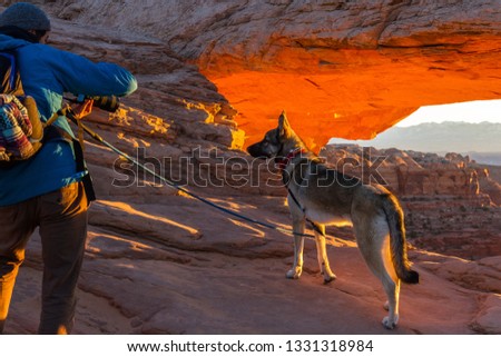 Man taking a picture of his dog with Mesa arch and the sunrise behind them