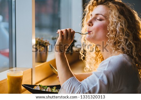 So tasty delicious yummy meal! Close up photo portrait of blond curly lady with closed eyes tasting healthy food from dish at cafe or restaurant in evening time. Royalty-Free Stock Photo #1331320313