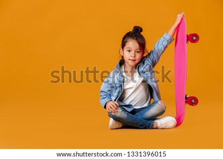 Stylish little child girl with skateboard in jeans clothes on orange background