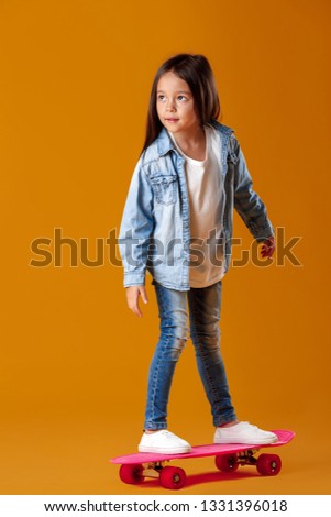 Stylish little child girl with skateboard in jeans clothes on orange background