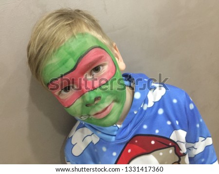 Blon boy on face green and red painted mask. Gray brown background.