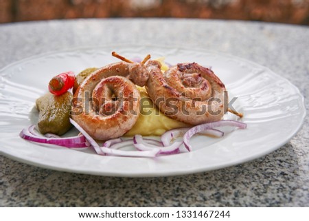 Czech traditional and seasonal food. Spiral fried white wine sausages with mashed potatoes, pickles from cucumber and hot pepper, chopped fresh red onion served on garden table outdoor on white plate 