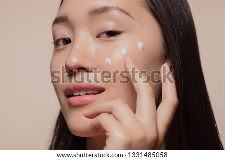 Close up of a young woman applying moisturizer to her face. Asian woman looking happy while following skincare regime. Royalty-Free Stock Photo #1331485058