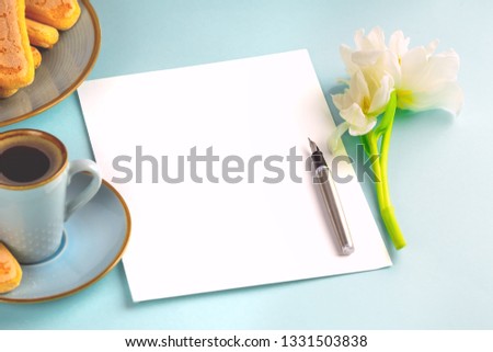 A sheet of white paper is empty on a blue background with an ink pen and a white tulip,there is also a blue cup with coffee on a saucer and a plate with Savoyardi biscuits. Place for text.