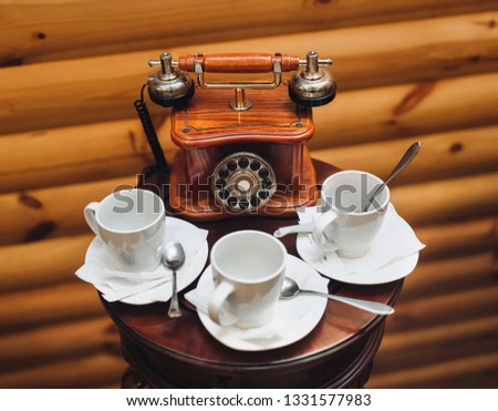 An old, antique, rarity telephone with a disk dialing set is standing on the table near empty white cups on a wooden background. Close up.