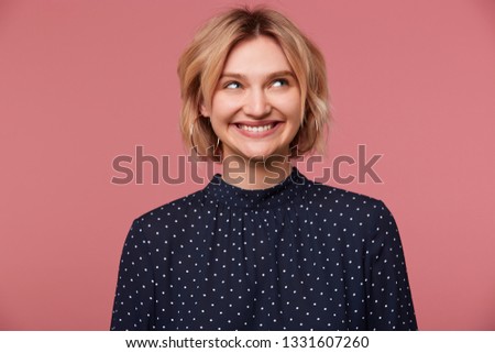 Happy thinking about boyfriend attractive beautiful blonde girl with short haircut looking at right upper corner touches her cheek dressed in blouse with polka dots, isolated over pink background