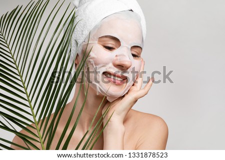 Woman with sheet facial mask and tropical leaf on light background