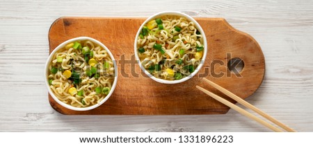 Instant noodles in paper cups on rustic wooden board over white wooden background, top view. Flat lay, from above, overhead. 