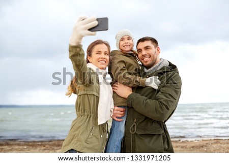 family, leisure and people concept - happy mother, father and little daughter taking selfie by smartphone on autumn beach