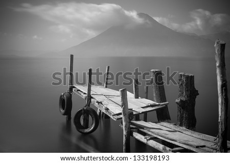 Old rickety wooden pier juts out into lake Atitlan (Guatemala) with volcano in background.  Black and White long exposure