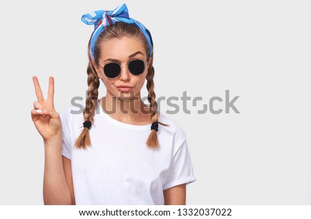 Studio portrait of cheerful blonde young woman wearing trendy sunglasses, white t-shirt and blue headband, making a duck face and showing peace sign. Student girl going crazy with braids hairstyle