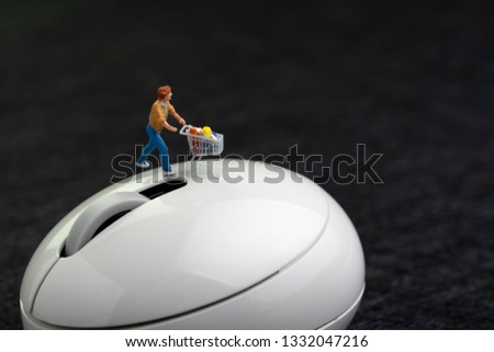 Miniature man with grocery in shopping cart or trolley walking on white computer mouse on dark black background with copy space using  online shopping or e-commerce website concept.