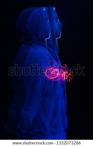 A male model with stubble wearing a hood up in a dark atmospheric future colour setting. Red and blue neon lighting gels. Blade runner neon futuristic shutter drag experimental light.