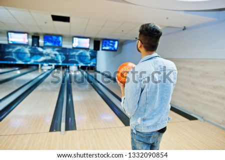 Back of stylish asian man in jeans jacket and glasses standing at bowling alley with ball at hand.