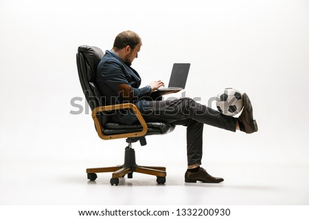 Businessman with football ball in office. Soccer freestyle. Concept of balance and agility in business. Manager perfoming tricks sitting on chair and working on laptop isolated on white studio Royalty-Free Stock Photo #1332200930