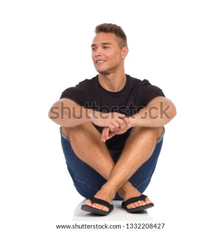 Young man in black shirt, jeans shorts and sandals, sitting relaxed on a floor and looking away and smiling. Full length studio shot isolated on white.