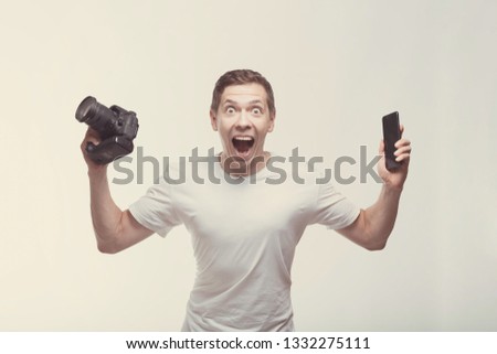 Emotional Man with camera isolated on light background. Young man holding camera and smartphone with victory screaming. Lifestyle, travel and technology concept. Glad boy in white t-shirt