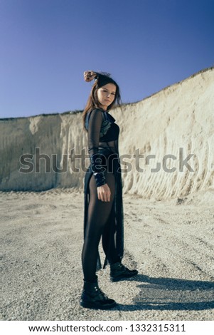 A young beautiful brunette girl in fashionable black clothes poses in sand quarry on sunny summer day. Sandpit. She raises one hand above her head. She looks at the camera.