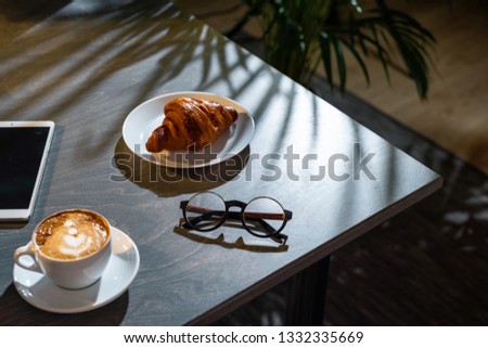 Close up of white coffee cup, croissant, glasses and pad with blank screen on dark wooden table with tropic plant on background.