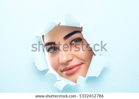 the face of a young beautiful girl with a bright make-up and puffy blue lips peers into a hole in blue paper.Fashion, beauty, make-up, cosmetics, hairstyle, beauty salon, boutique, discounts, sales.