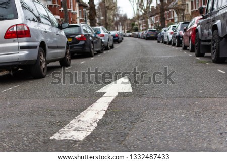 A residential street where parked cars line the sides of the street. The angle is low and there is a white arrow painted onto the road, pointing forward, indicating a one way road.