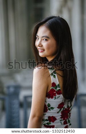 Close up portrait head shot of a young and attractive Chinese Asian girl in a white and red summer floral dress against a grey courtroom in the city. She is petite and pretty and is smiling. 