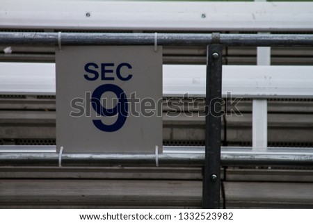 Offset view of the section number (Sec 9) at a sports venue. The section number sign is white with letters in navy blue Sec 9. The sign is wire tied between two galvanized aluminum pipes.