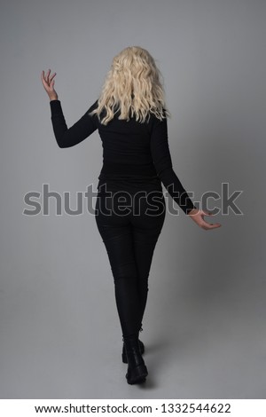 full length portrait of a blonde girl wearing  modern black jacket and pants, standing pose on grey studio background.