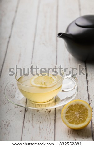 Homemade organic cough and cold relief, made of  a glass cup of hot drink with a slice of lemon in it. There is a black teapot in the background, and there is a halved lemon in the foreground.