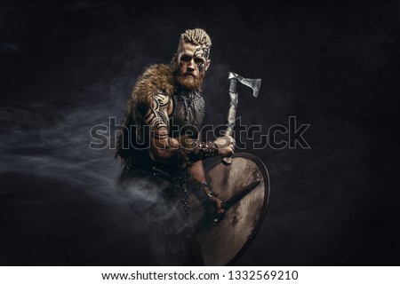 Medieval warrior berserk Viking with tattoo and in skin with axes attacks enemy. Concept historical photo Royalty-Free Stock Photo #1332569210