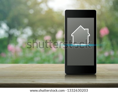 House flat icon with copy space on modern smart mobile phone screen on wooden table over blur pink flower and tree in garden, Businesss real estate online concept