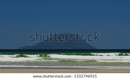 in the foreground the sand of the beach and the waves breaking in the sea of ​​green color, in the background distant island covered by a slight fog under a blue sky, north coast of sao paulo, Brazil