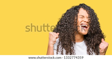 Young beautiful woman with curly hair wearing white t-shirt very happy and excited doing winner gesture with arms raised, smiling and screaming for success. Celebration concept.