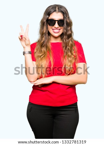 Young beautiful woman wearing red t-shirt and sunglasses smiling with happy face winking at the camera doing victory sign. Number two.