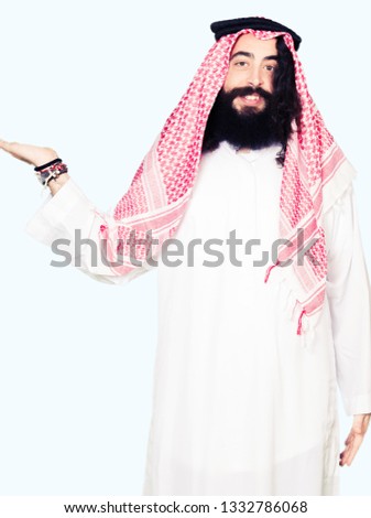 Arabian business man with long hair wearing traditional keffiyeh scarf smiling cheerful presenting and pointing with palm of hand looking at the camera.