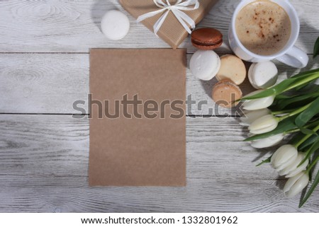 Bunch of white tulips, gifts,  macaroons, cup of coffee and sheet of paper for message on white wooden table. Greeting concept. Spring menu, women or mothers day, cakes, cappuccino