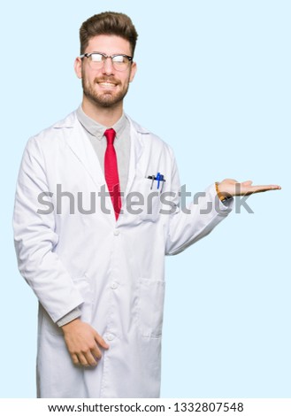 Young handsome scientist man wearing glasses smiling cheerful presenting and pointing with palm of hand looking at the camera.