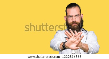 Young blond doctor man with beard wearing medical coat Rejection expression crossing arms and palms doing negative sign, angry face