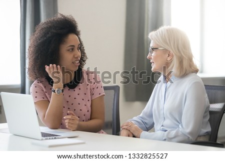 Diverse old and young female colleagues talking at work, african and caucasian business women sitting together in office having friendly conversation, mentor intern discussing planning shared project Royalty-Free Stock Photo #1332825527