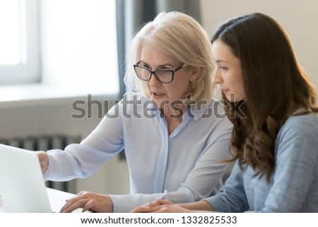 Serious old female mentor teacher coach teaching intern or student computer work pointing at laptop, mature executive manager explaining online project to young employee learning new skills in office Royalty-Free Stock Photo #1332825533