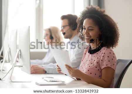 Smiling african american businesswoman call center operator agent wearing headset holding papers reading clients contacts working in customer service support helpdesk business office with colleagues Royalty-Free Stock Photo #1332843932