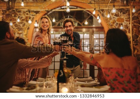 Host couple toasting drinks with guest at dinner party. Young cheerful people celebrating with drinks at party.