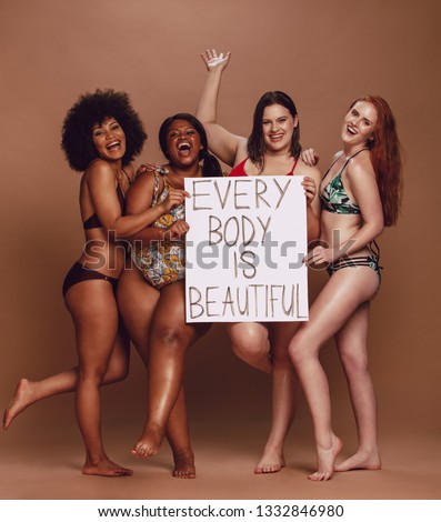 Cheerful group of women holding a every body is beautiful signboard over brown background. Multiracial females of different figure and size looking excited together.