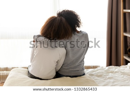 Rear back view black mother and daughter embrace sitting on bed at home, older sister consoling younger teen, girl suffers from unrequited love share secrets trustworthy person relative people concept Royalty-Free Stock Photo #1332865433