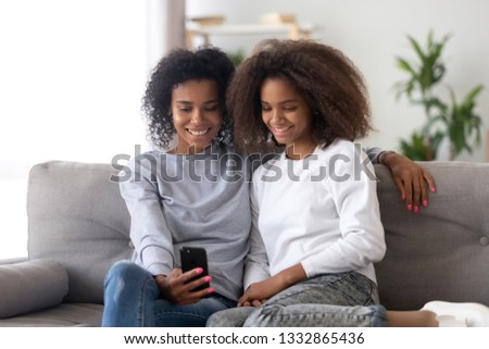 Different ages black african sisters having fun using application holding smartphone making video call with friends. Young mother spending free time with adolescent daughter take selfie photo at home