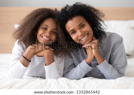 Happy african different ages sisters lying on bed at home or hotel bedroom smiling posing looking at camera, young mom spending free time with teenage daughter, lazy weekend leisure activities concept Royalty-Free Stock Photo #1332865442